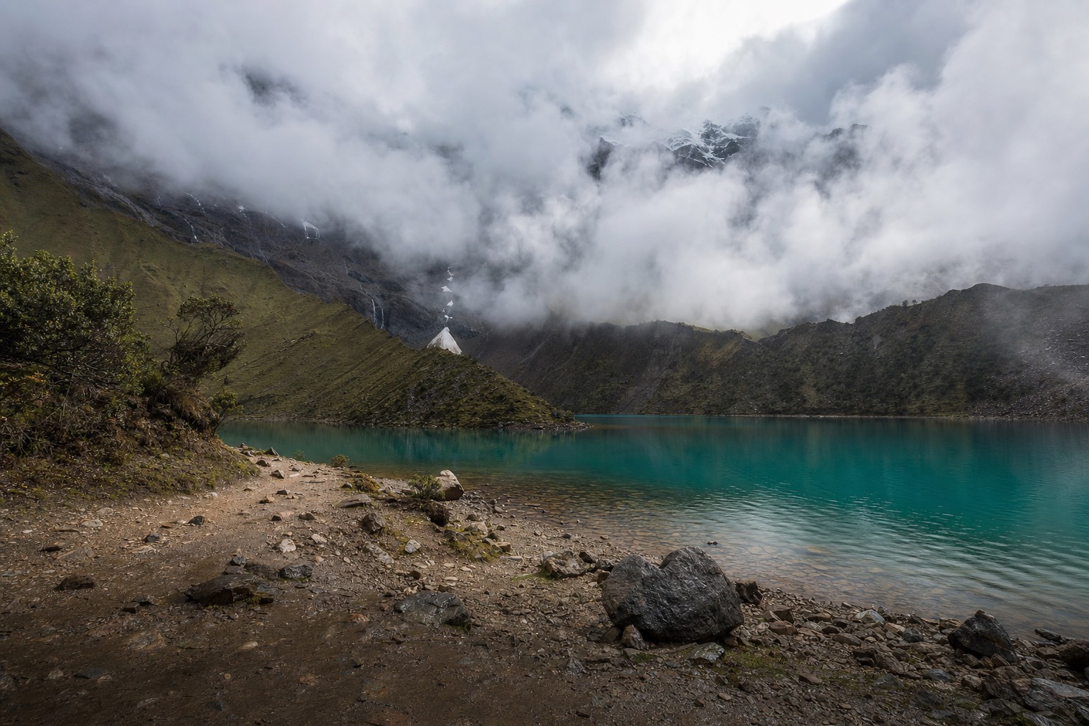 Mist and low cloud drifting over the turquoise water of Humantay Lake with a wet rocky trail in the foreground