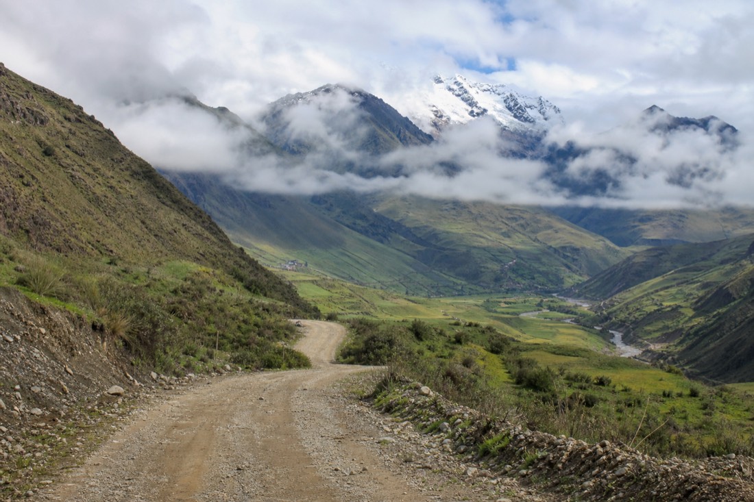 Unpaved mountain road descending through a broad green Andean valley with distant snow peaks under morning cloud, no people or vehicles