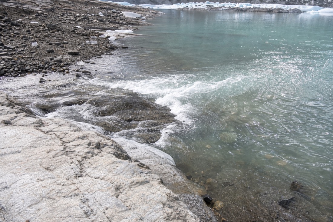 Icy meltwater inlet where a glacier stream enters a turquoise high-altitude lake, pale rock and thin ice at the edge, no people