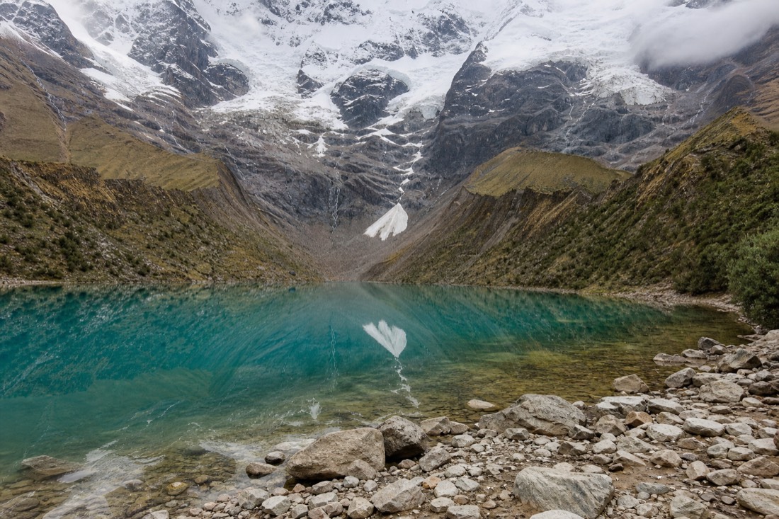 Still turquoise water of Humantay Lake at the shore with the glacier and Humantay peak reflected, loose rock in the foreground
