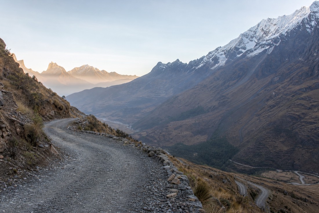 Narrow gravel switchback road climbing a steep Andean slope toward Soraypampa at dawn with the Salkantay valley below and no vehicles in frame
