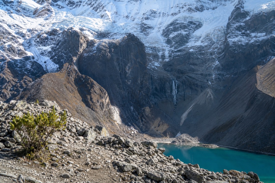 Rocky moraine ridge near the top of the Humantay trail with the edge of the turquoise lake and a hanging glacier beyond in morning light, no people