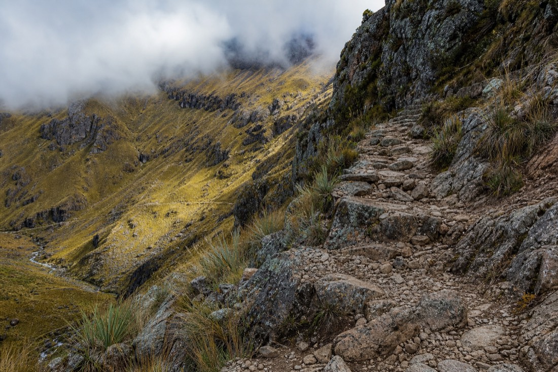 Close view of loose rocky switchback steps climbing a steep Andean slope above a grassy valley with no people