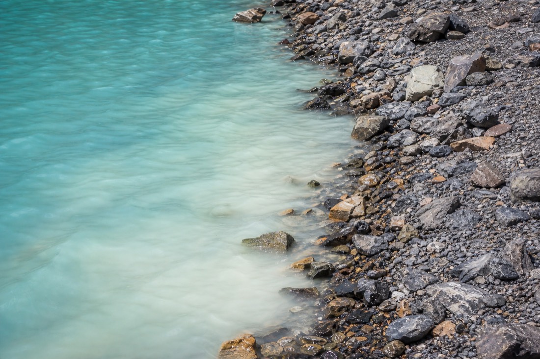 Close view of pale turquoise glacial meltwater meeting grey moraine gravel at the edge of Humantay Lake, no people